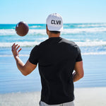Man tossing an american football at the beach wearing a Black Clover Live Lucky CLVR hat in white.