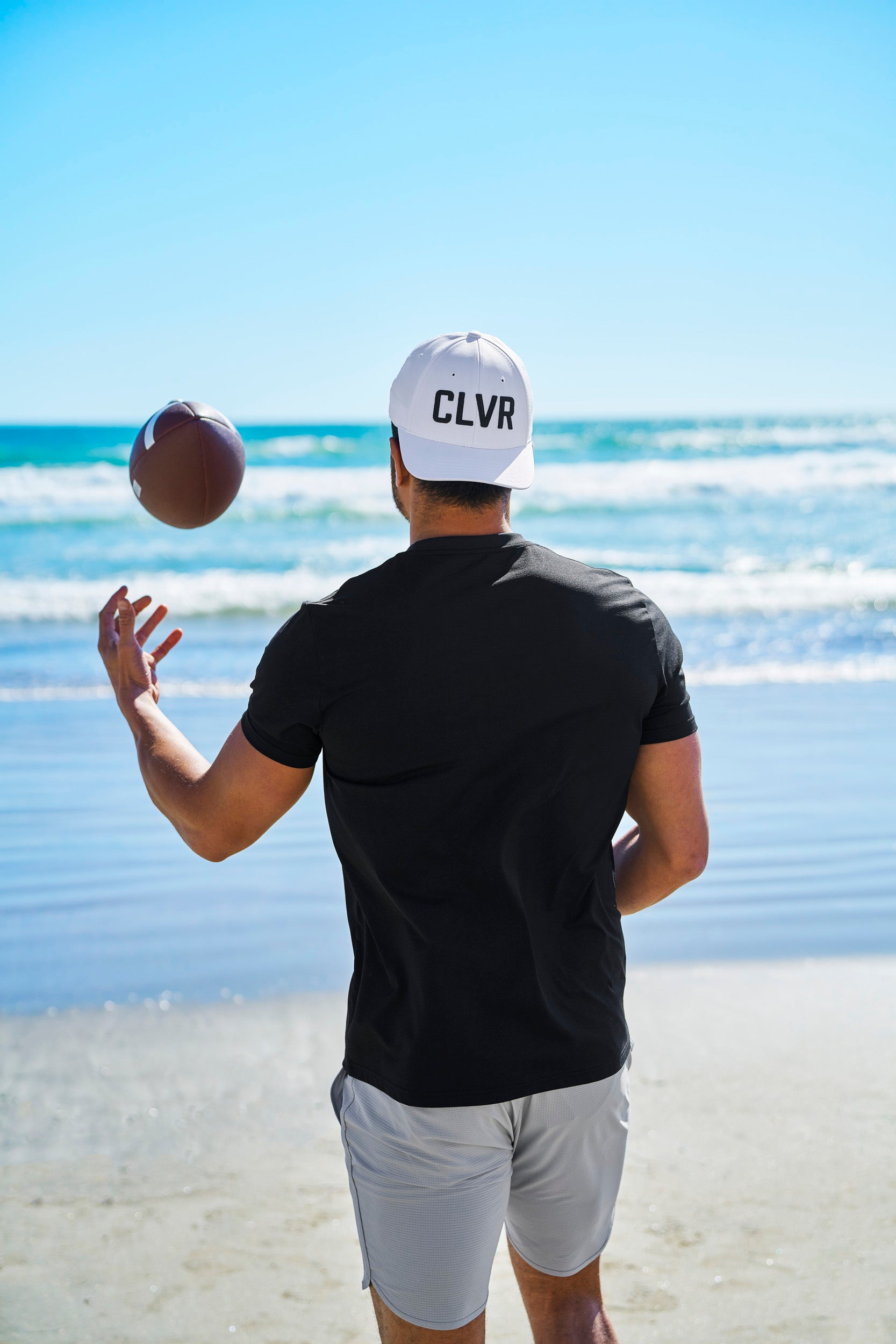 Man tossing an american football at the beach wearing a Black Clover Live Lucky CLVR hat in white.